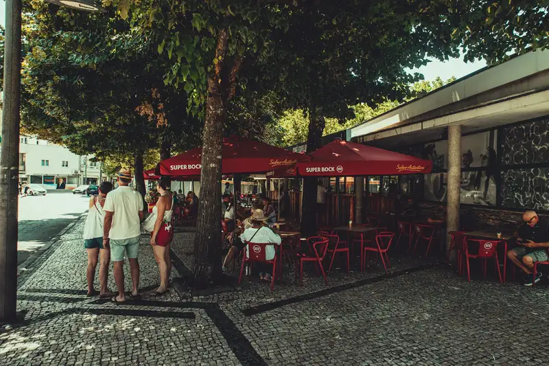 Outdoor caf茅 with red umbrellas, people relaxing under trees. Summer day in urban setting.